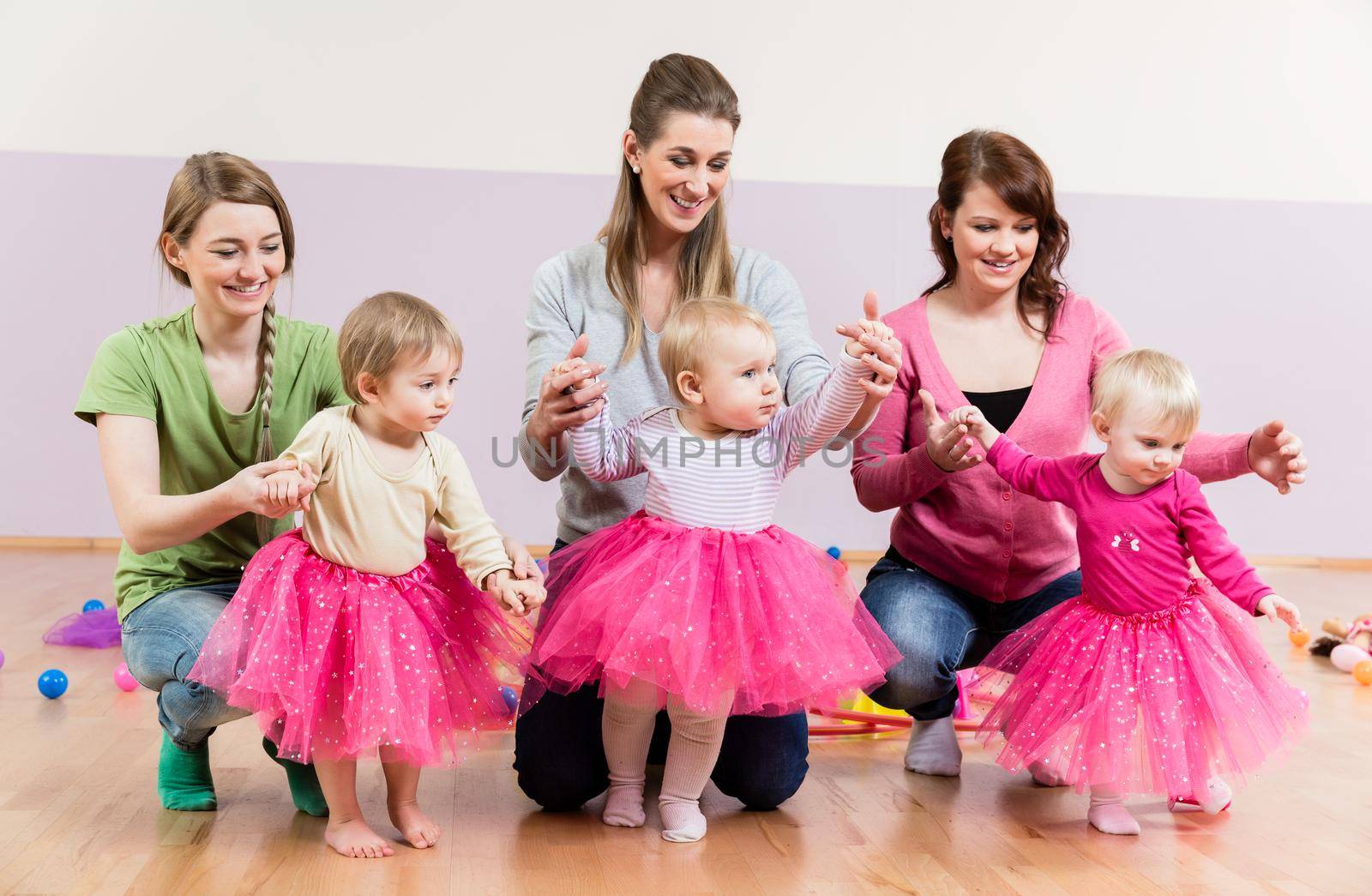 Three baby girls in pink skirts learning to walk with help of their mothers