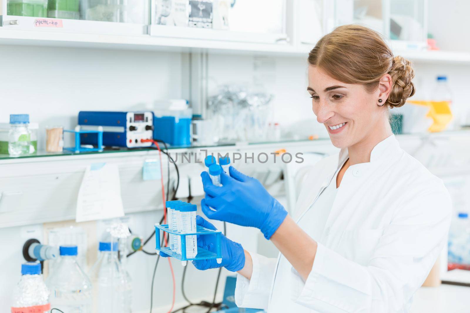 Cheerful woman researcher looking at test-tubes in laboratory while holding stand
