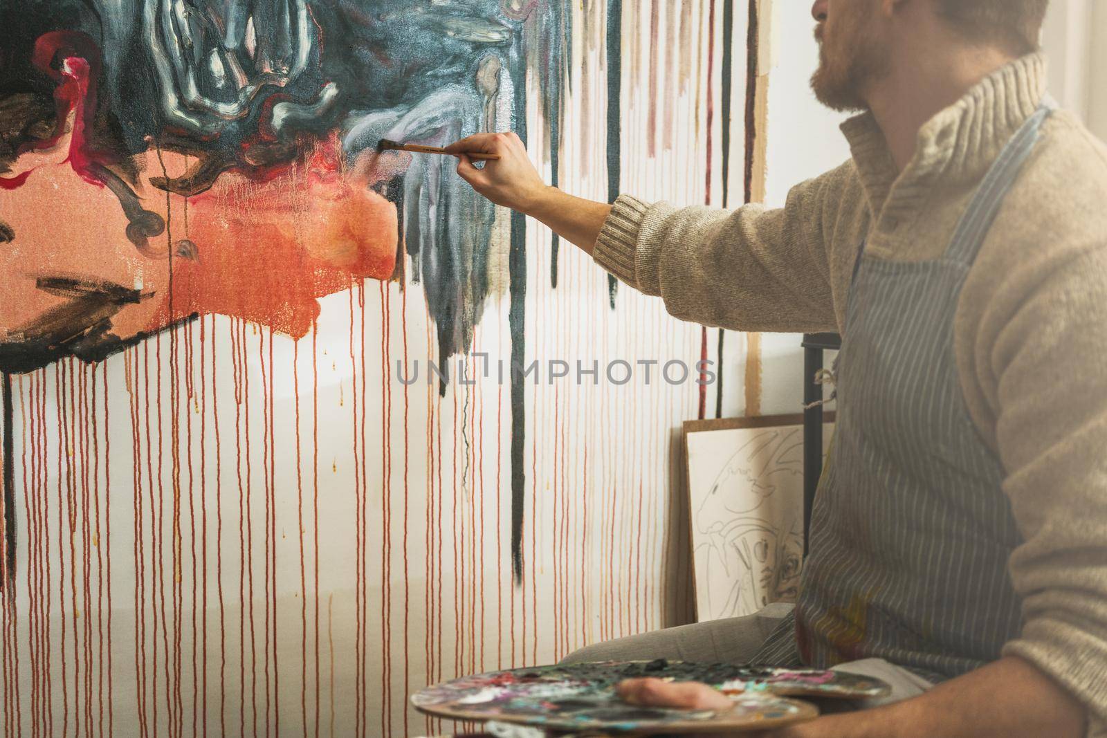 Confident male artist holding palette and paint abstract painting with paint brush on board in art studio