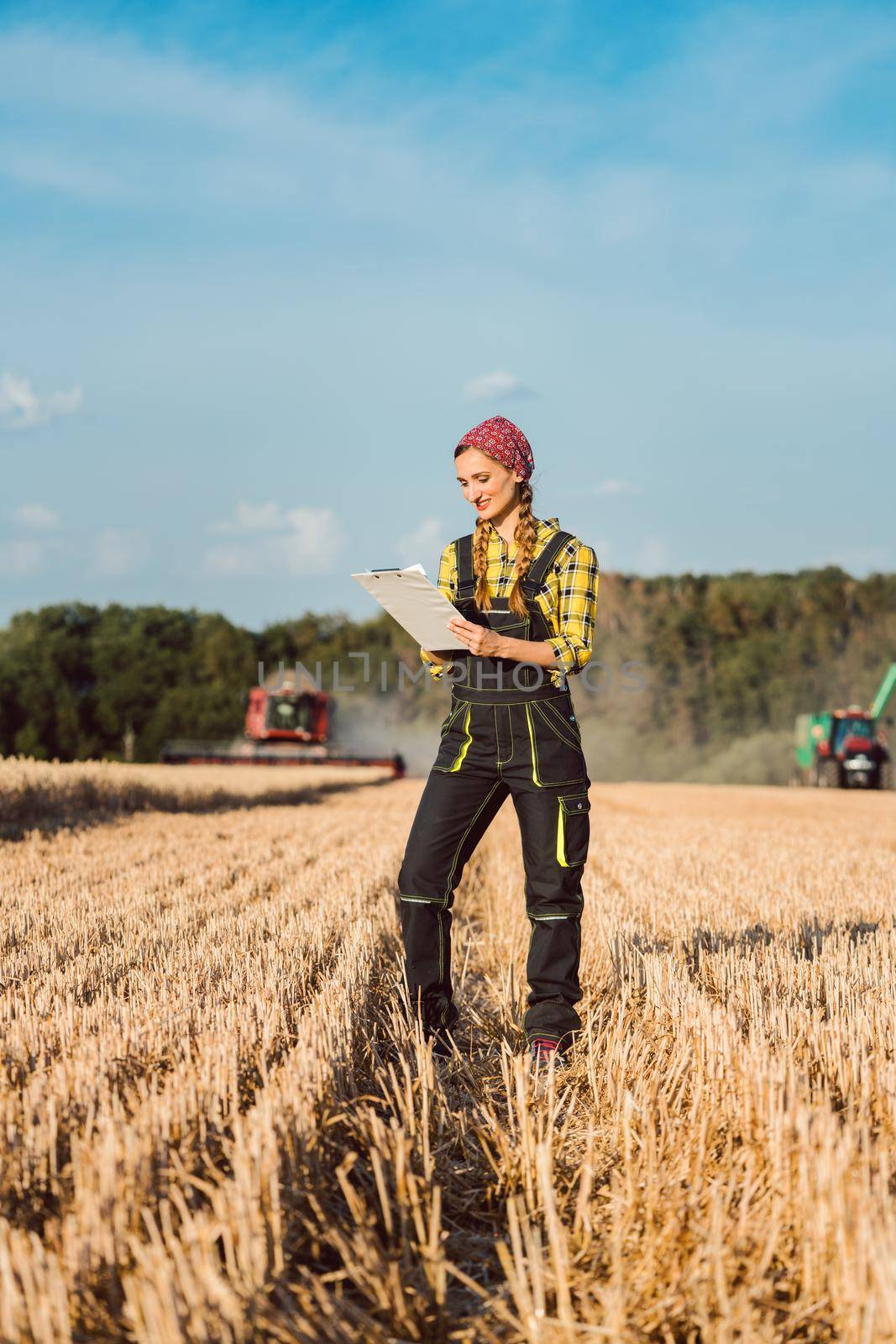 Farmer woman monitoring business progress of the harvest with agricultural machinery in the background