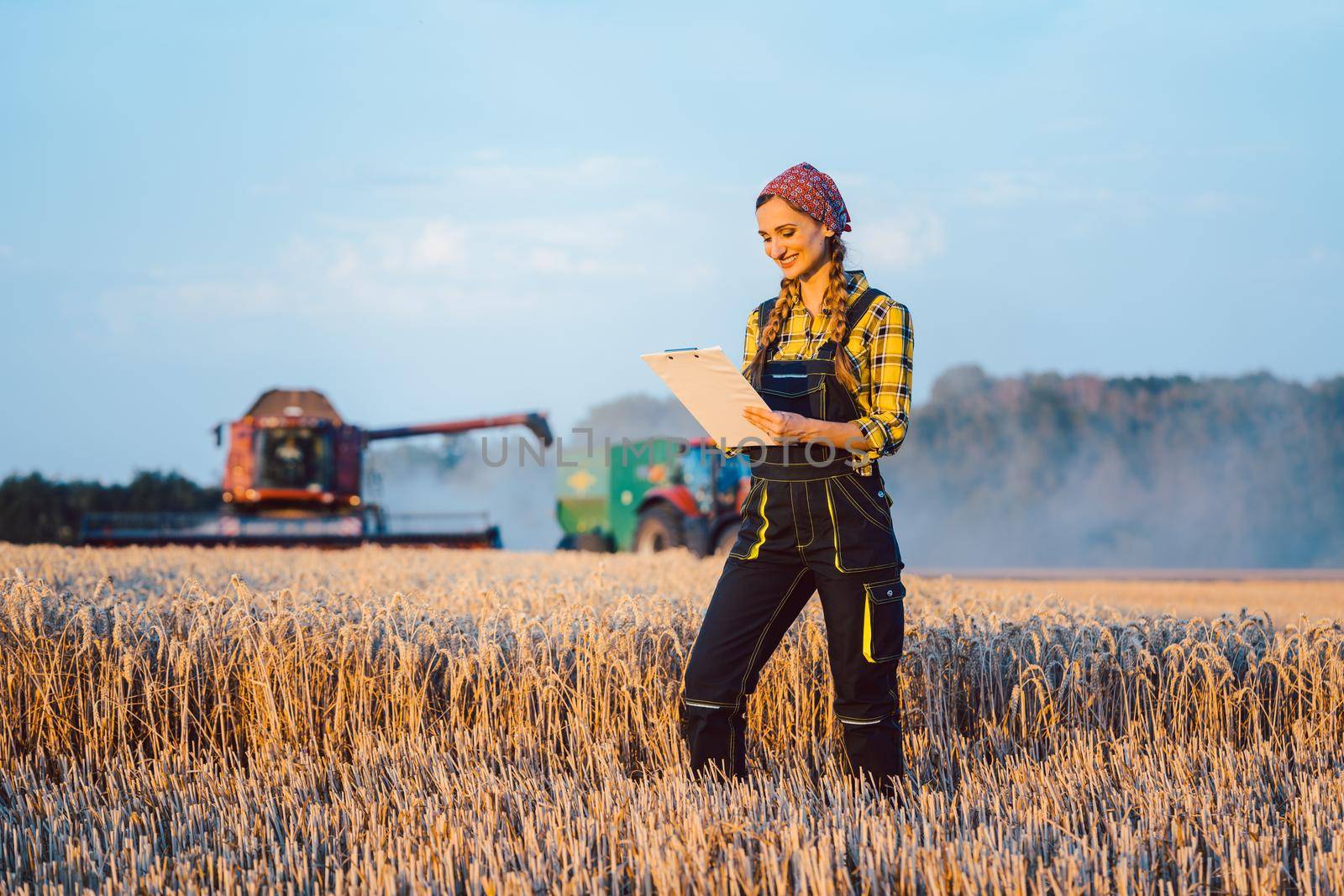 Farmer woman with clipboard on field, harvest going on, combine harvester in the background