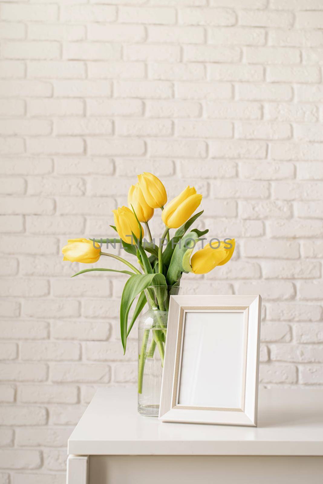 Bouquet of yellow tulips in a glass vase and blank photo frame on a white brick wall background. Mock up design