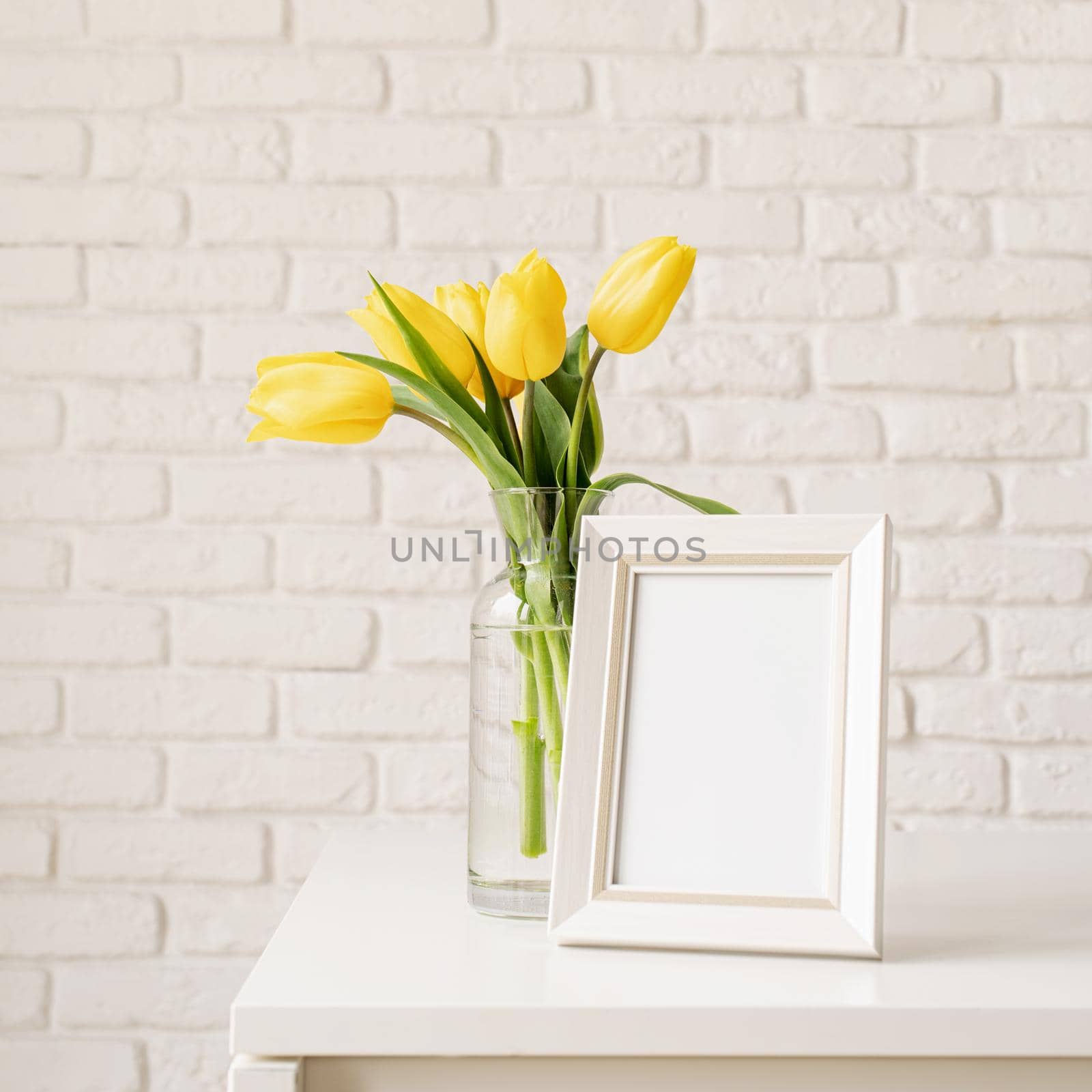 Bouquet of yellow tulips in a glass vase and blank photo frame on a white brick wall background. Mock up design