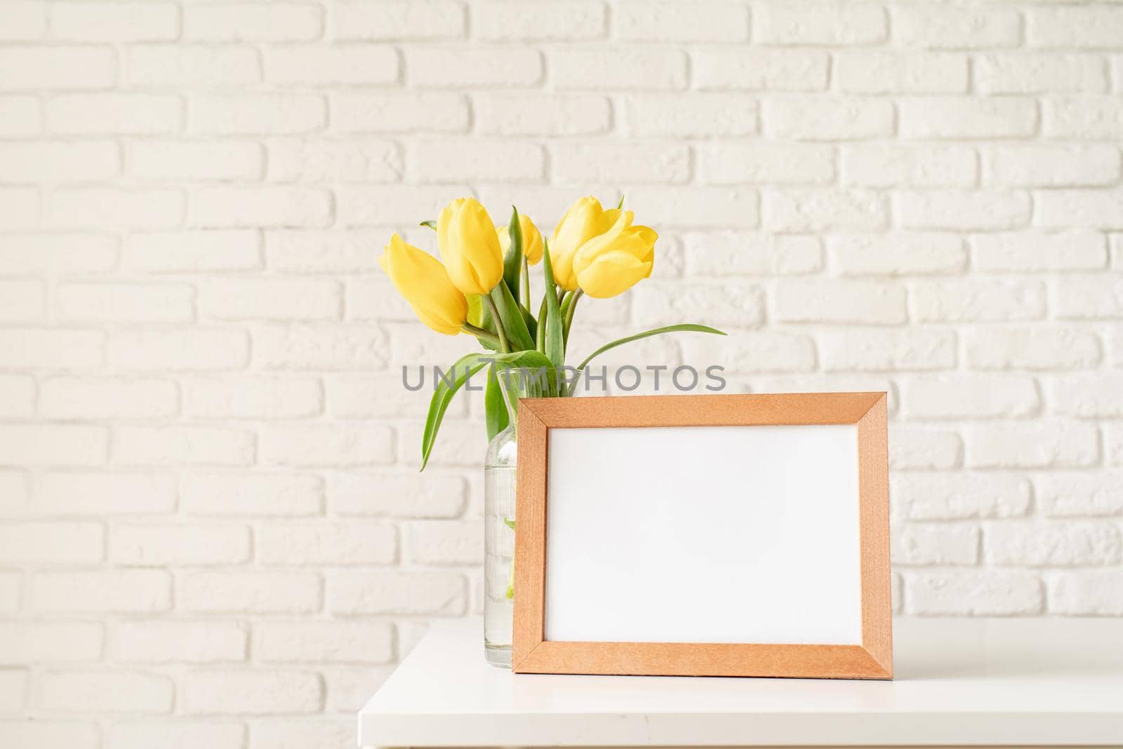 Bouquet of yellow tulips in a glass vase and blank photo frame on a white brick wall background. Mock up design