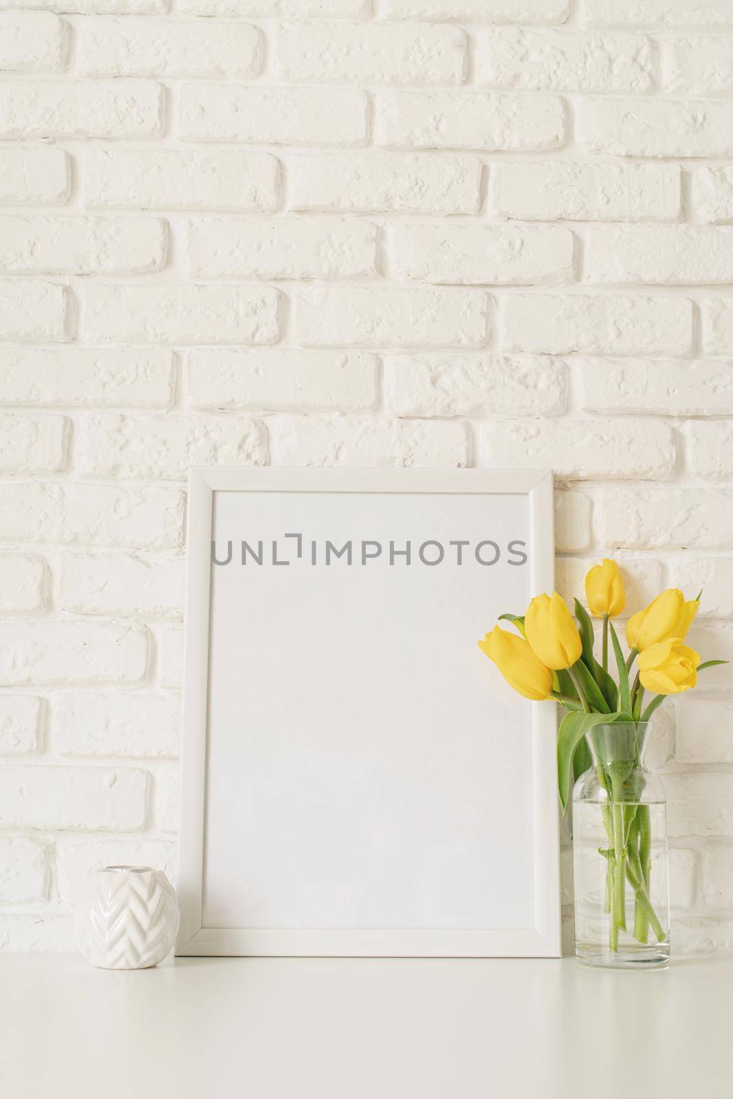 Bouquet of yellow tulips in a glass vase and blank photo frame on a white brick wall background. Mock up design