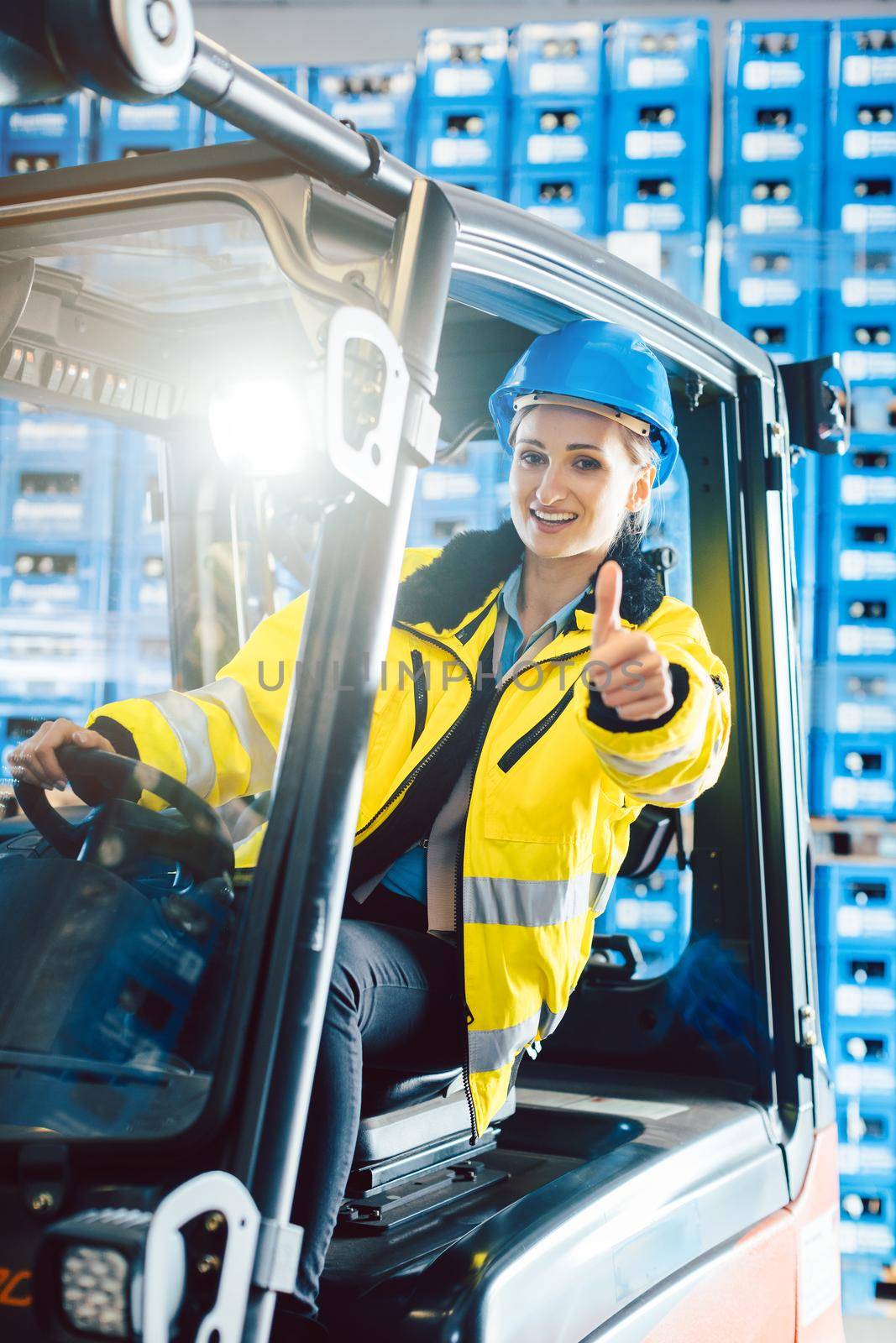 Worker woman showing thumbs up in logistics delivery center sitting in a forklift