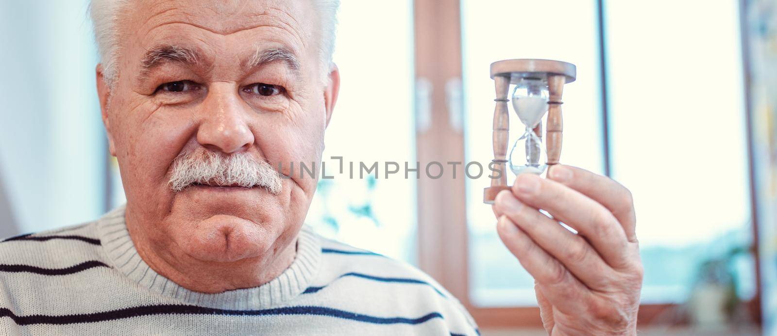 Senior man holding hourglass in retirement home, symbol for limited life by Kzenon