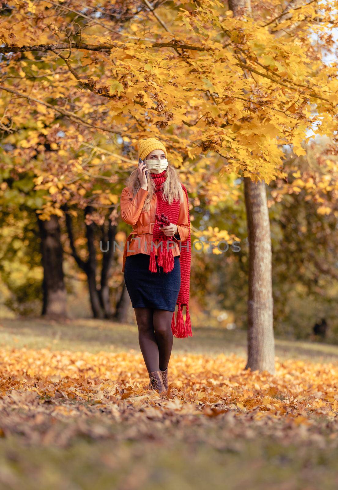 Woman using her phone having walk in fall park during coronavirus crisis amidst colorful foliage