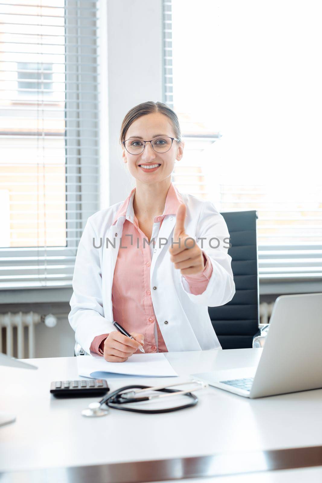 Doctor woman giving thumbs-up into the camera sitting on desk in her office