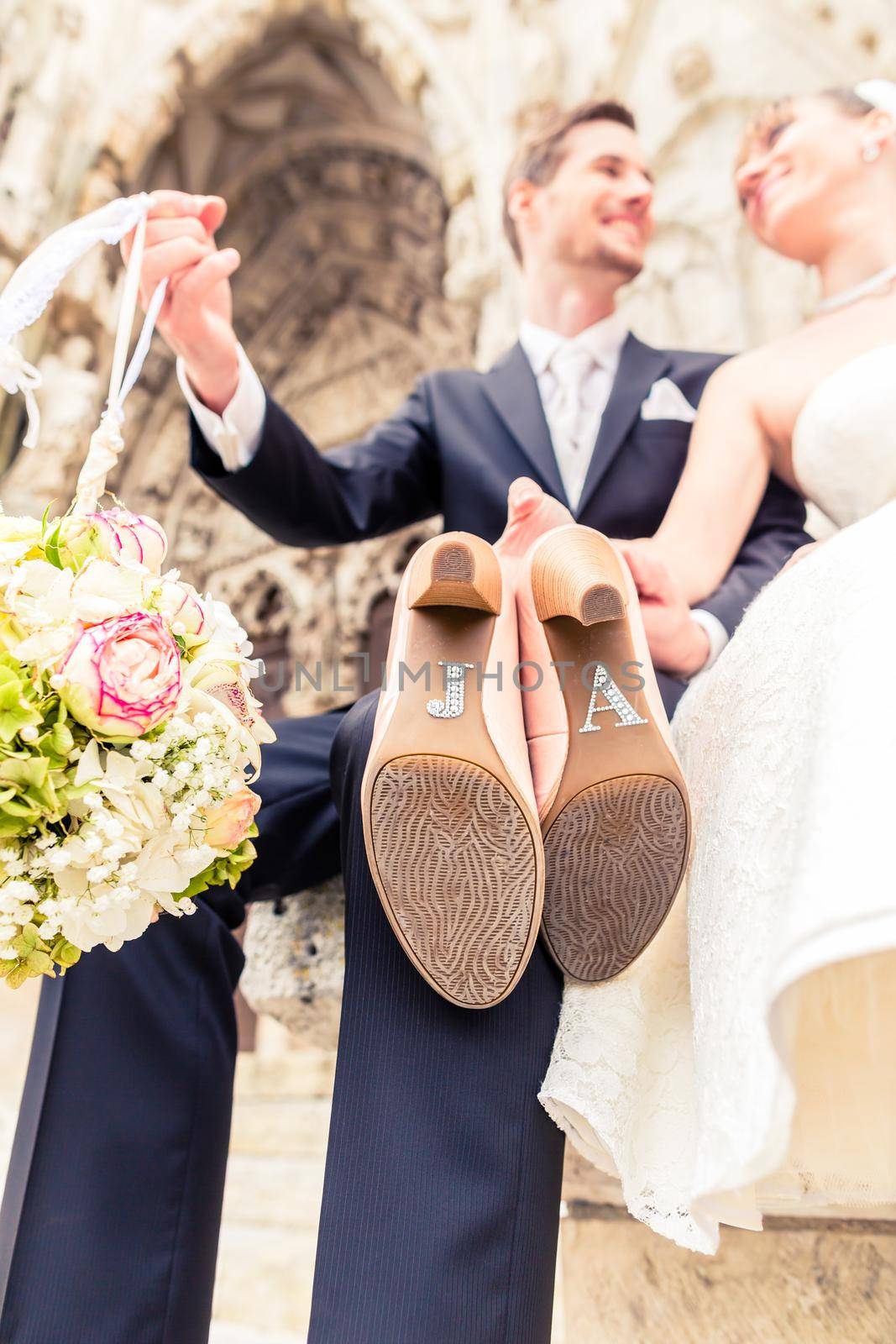 Smiling newlywed couple holding bouquet ball and high heels sitting outside the church