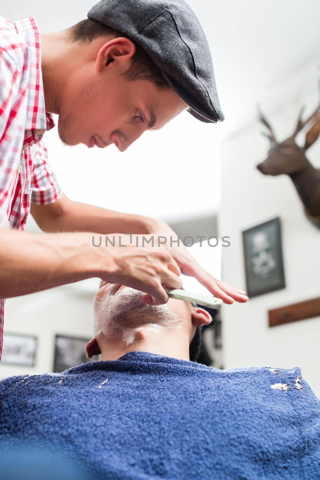 Close-up of young male barber shaving with straight razor in barbershop