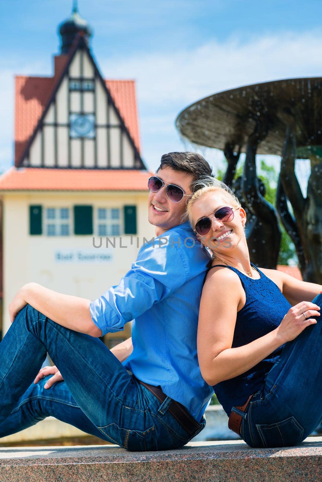 Happy young couple wearing sunglasses sitting back to back near the fountain