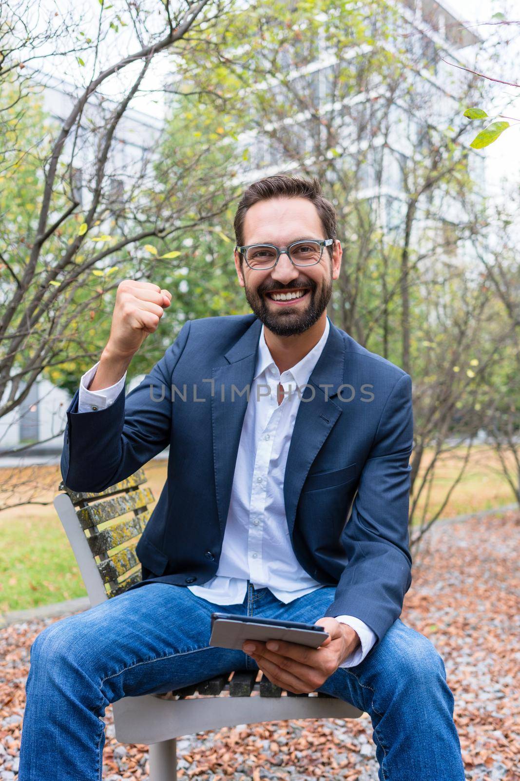 Portrait of successful businessman sitting on park bench holding digital tablet clenching his fist