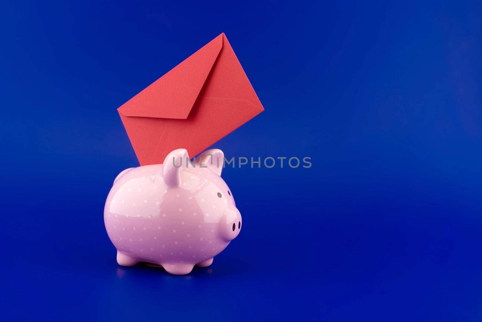 Red envelope being inserted into a pink piggy bank over blue background with copy space