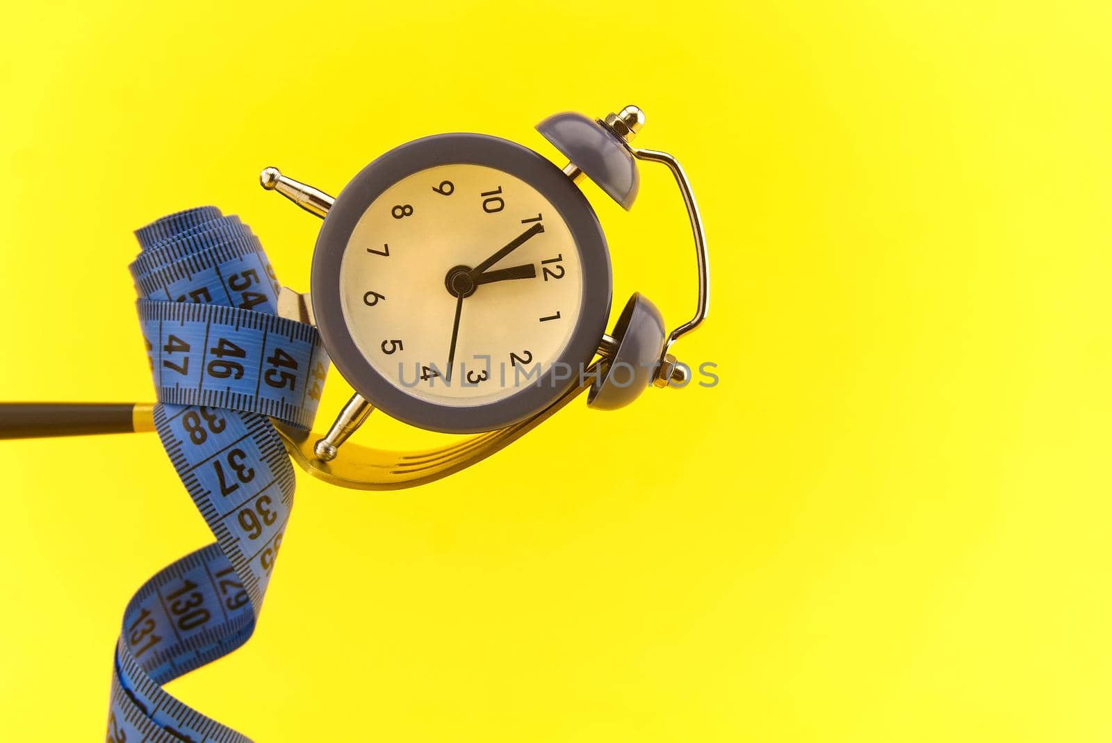 Alarm clock on golden fork wrapped with measuring tape above yellow background, weight loss or diet concept with copy space