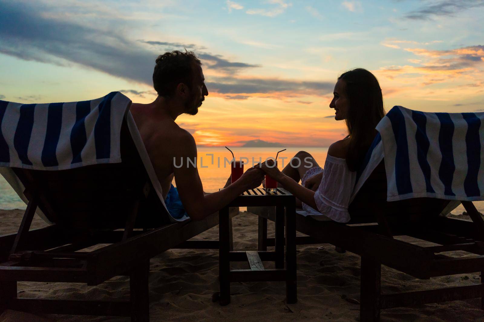 Rear view of a young romantic couple in love, sitting on wooden chairs while drinking cocktails on a tropical beach at sunset during vacation or honeymoon in Indonesia