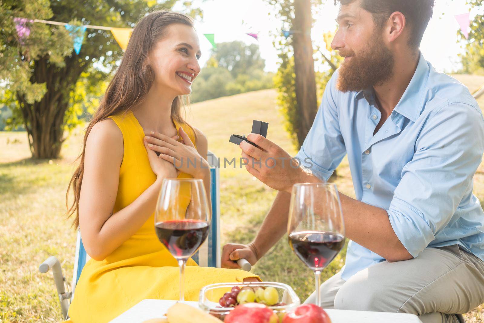 Young handsome man sitting on bended knee while showing to his beautiful girlfriend an engagement ring during romantic picnic with red wine and fresh fruits