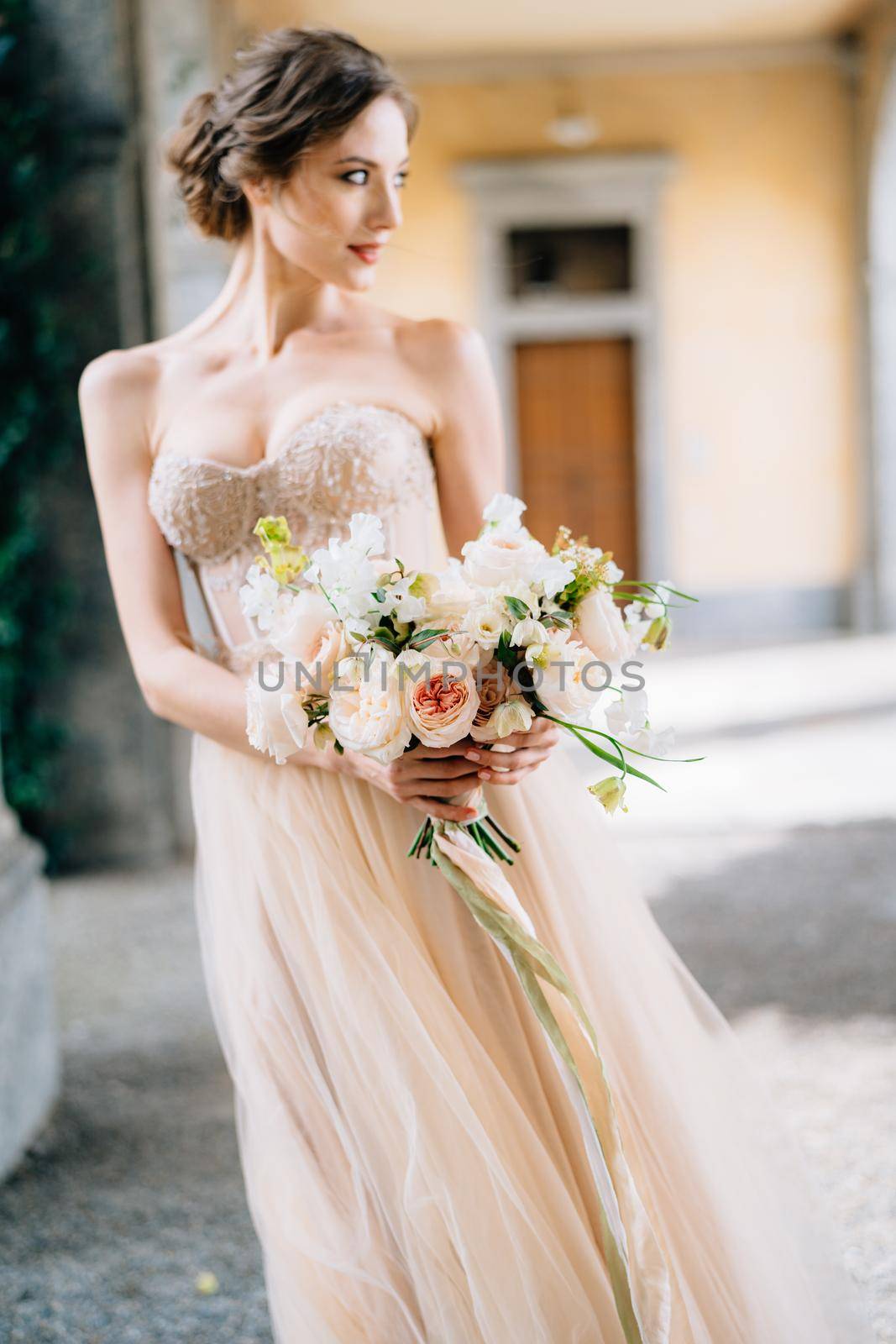 Bride in a beautiful dress with a bouquet of pink flowers stands in the vaulted hall with her head turned. Lake Como, Italy. Close up. High quality photo