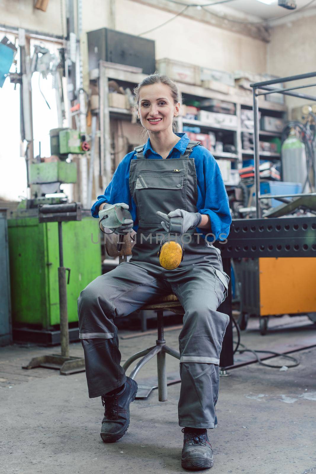 Female mechanic sitting in metal workshop looking and smiling into camera