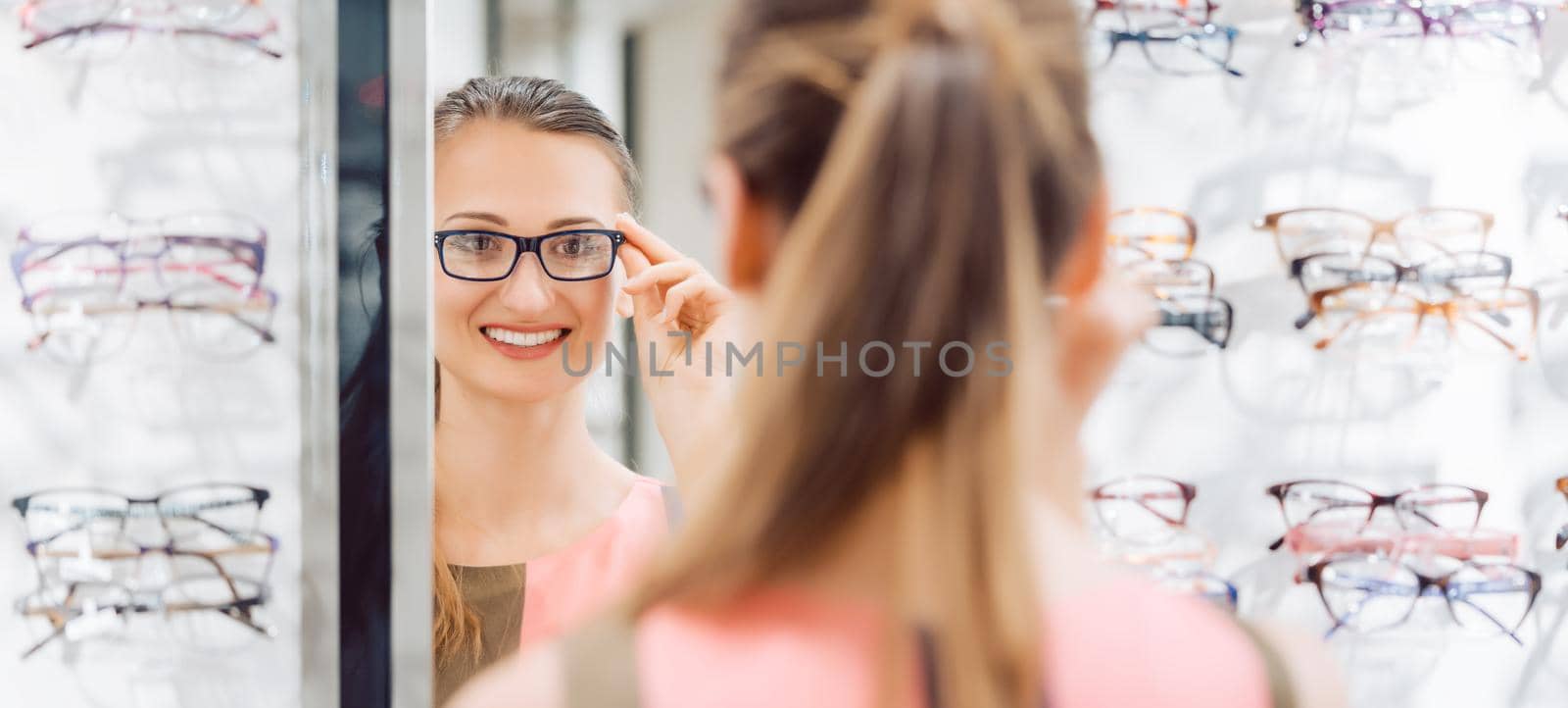 Young woman trying fashionable glasses in optometrist store by Kzenon