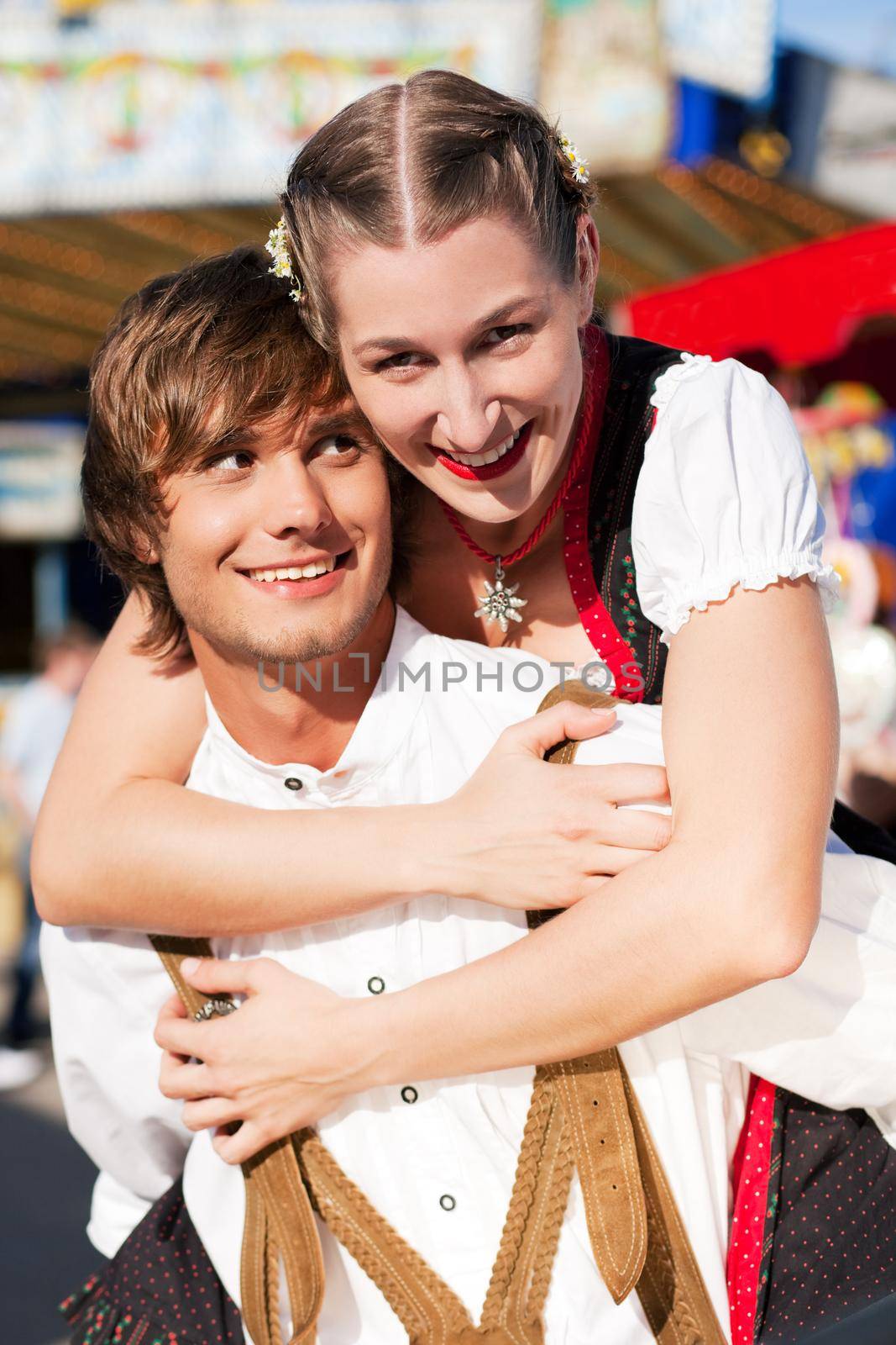 Young and beautiful couple in traditional Bavarian Tracht - Dirndl and Lederhosen - embracing each other on a fair like a Dult or the Oktoberfest; both are standing in front of a typical booth