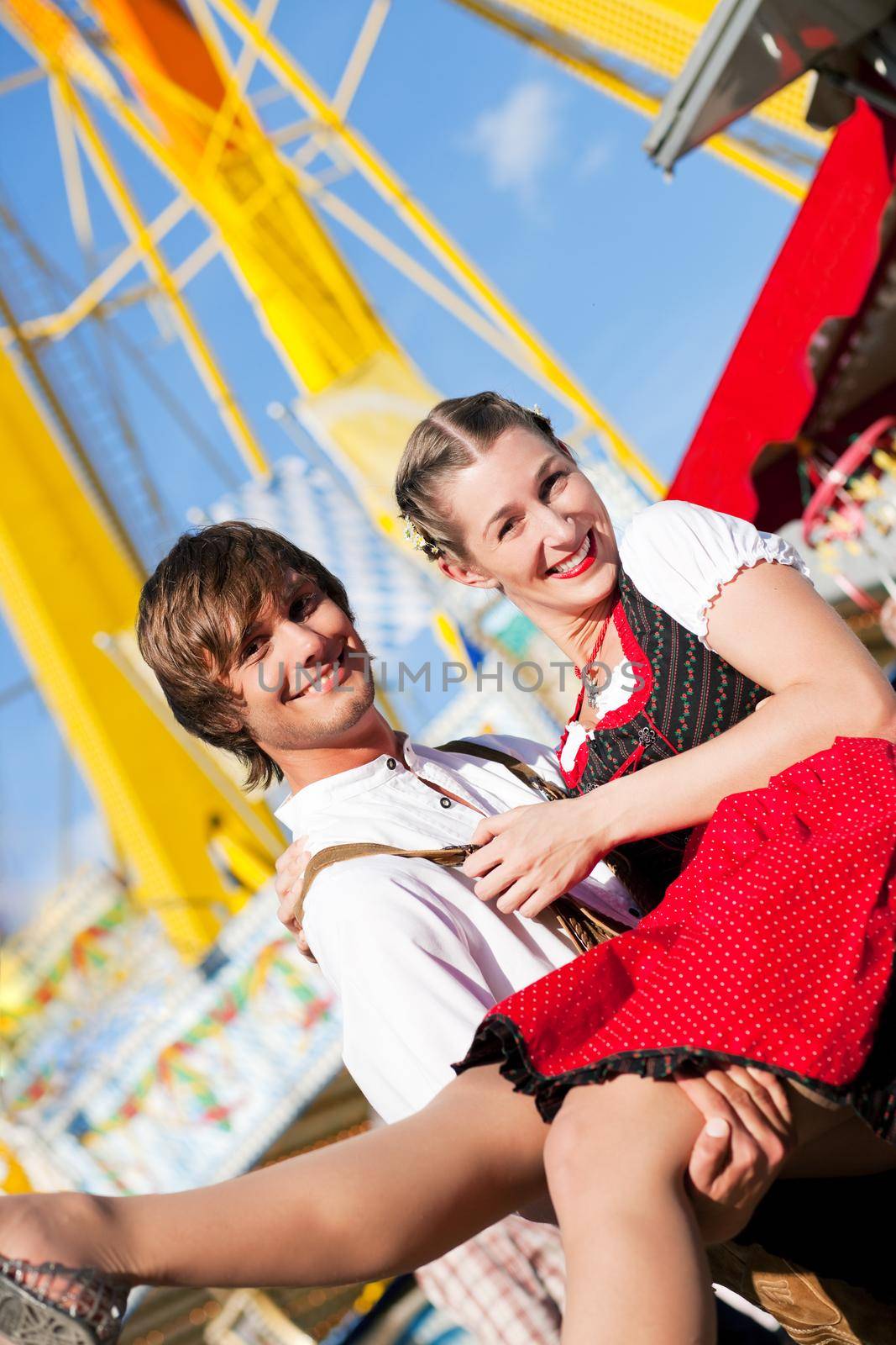 Young and beautiful couple in traditional Bavarian Tracht - Dirndl and Lederhosen - embracing each other on a fair like a Dult or the Oktoberfest; both are standing in front of a typical booth