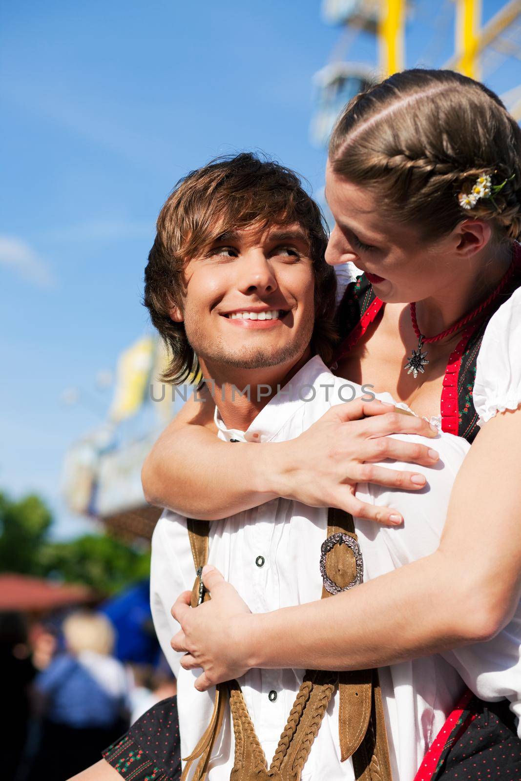 Young and beautiful couple in traditional Bavarian Tracht - Dirndl and Lederhosen - embracing each other on a fair like a Dult or the Oktoberfest; both are standing in front of a typical booth