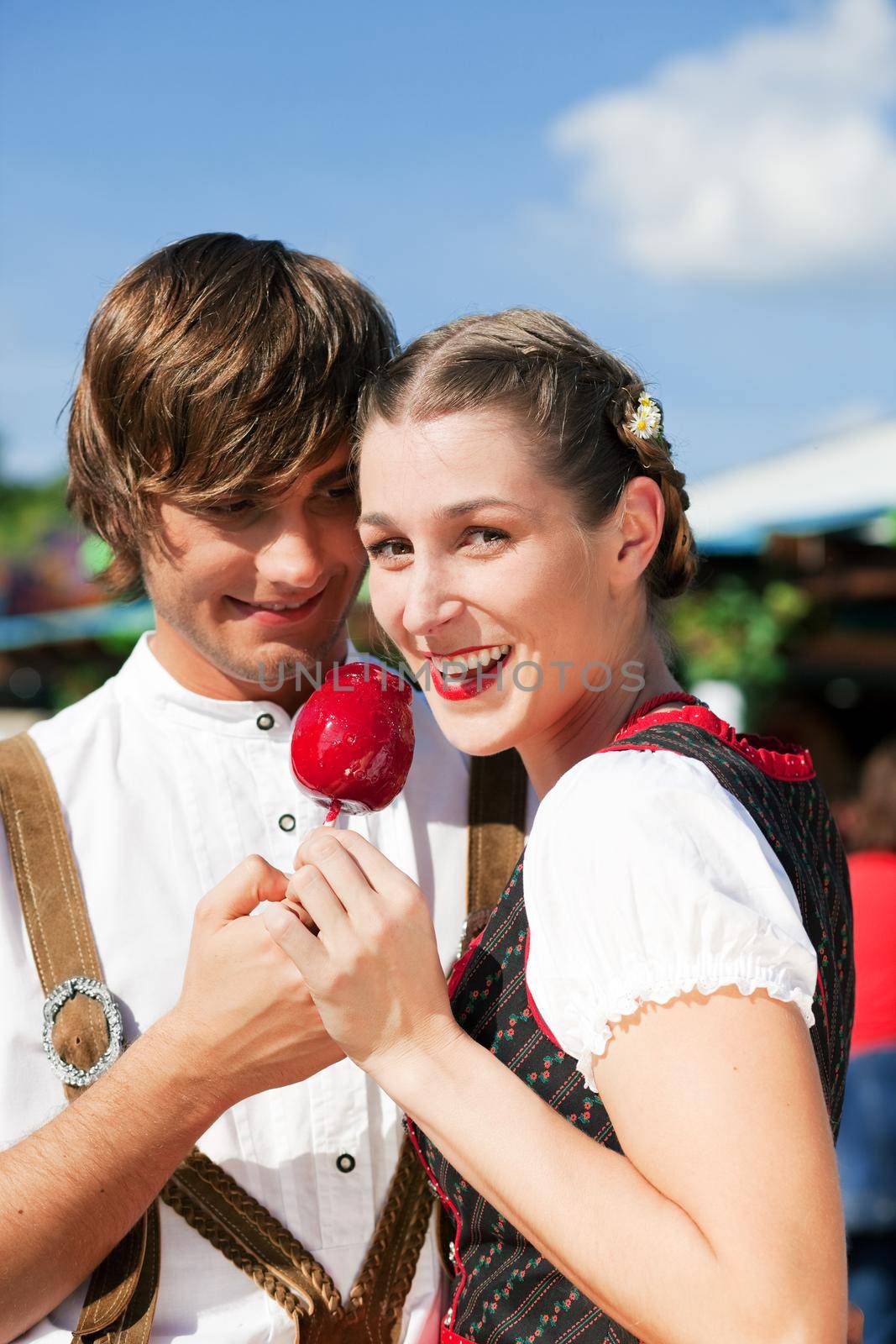 Young and beautiful couple in traditional Bavarian Tracht - Dirndl and Lederhosen - embracing each other on a fair like a Dult or the Oktoberfest eating traditional sugar apple