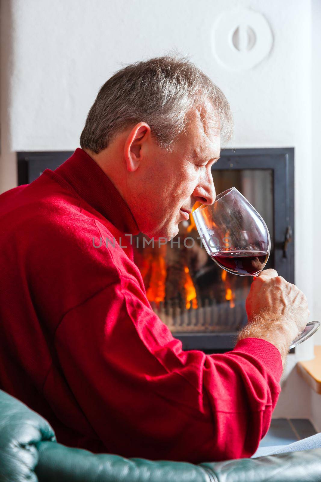 Mature man drinking a glass of red wine in front of his fireplace