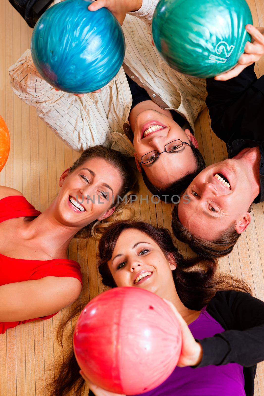 Group of four friends lying in a bowling alley having fun, holding their bowling balls above them