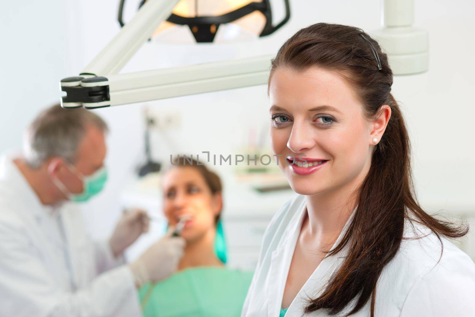 Dentists in her surgery looking at the viewer, in the background her colleague is giving a female patient a treatment
