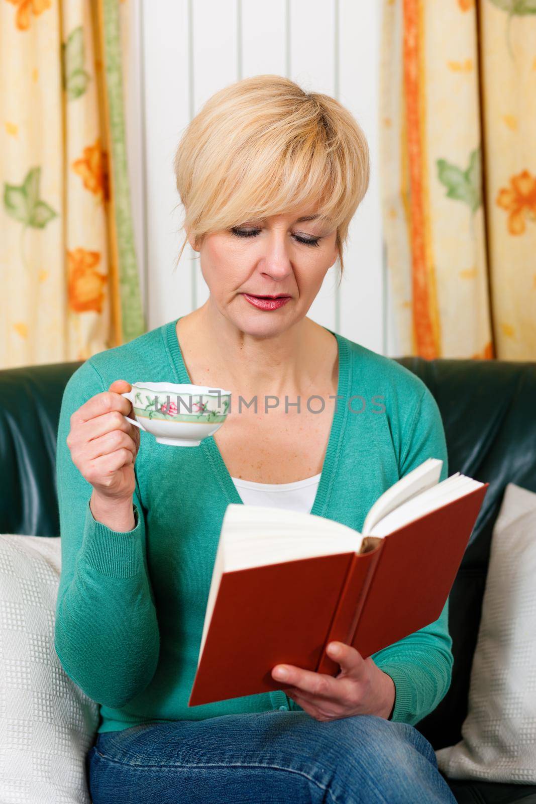 Mature woman is reading a book and is drinking a cup of coffee