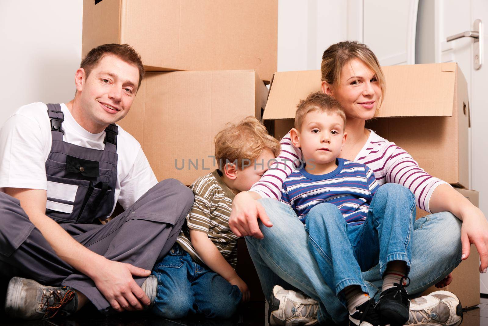 Family moving in their new house. They are sitting in front of a stack of moving boxes being happy. Father is dressed in a way that can also represent a mover