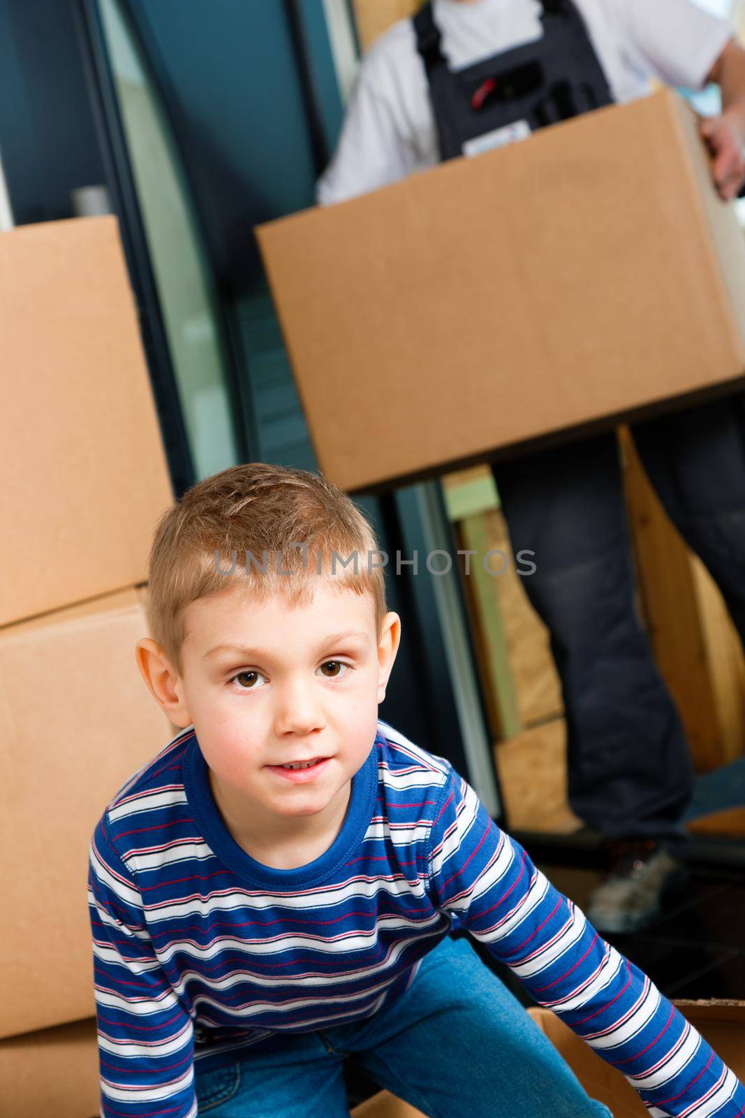 Family moving in their new home. The son is playing with a moving box. In the background the father - or a mover (only legs to be seen) is carrying boxes inside the building