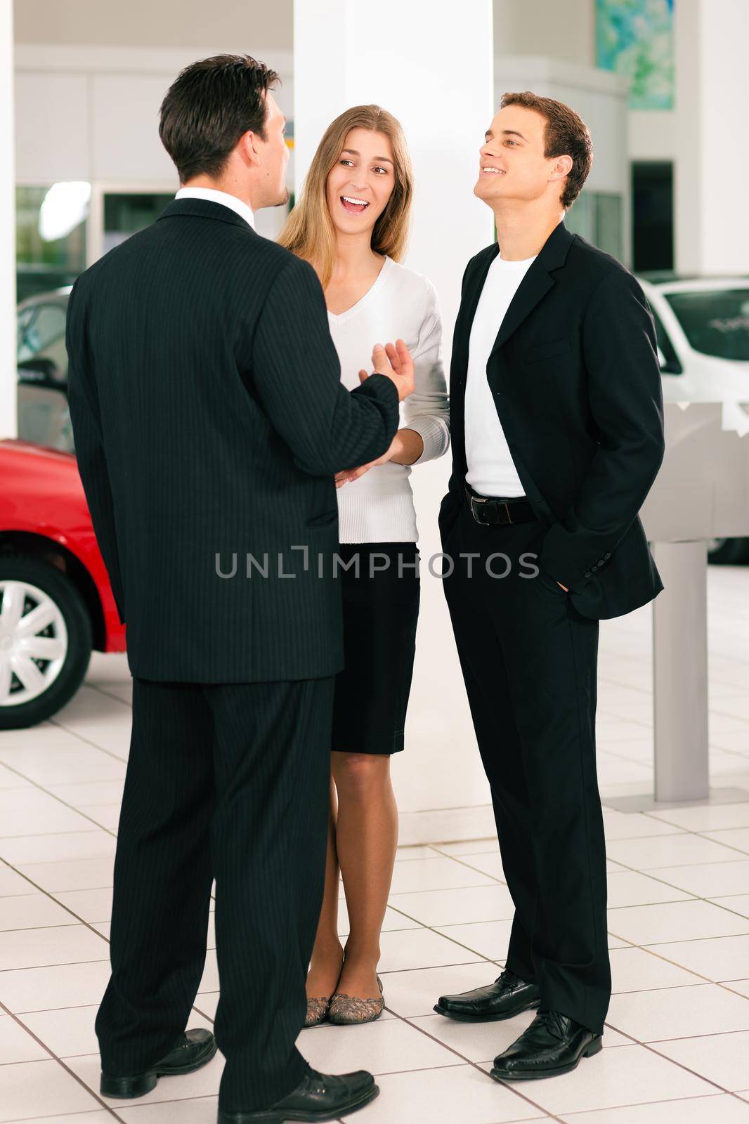 Sales situation in a car dealership, the dealer is talking to a young couple, there are cars standing in the background