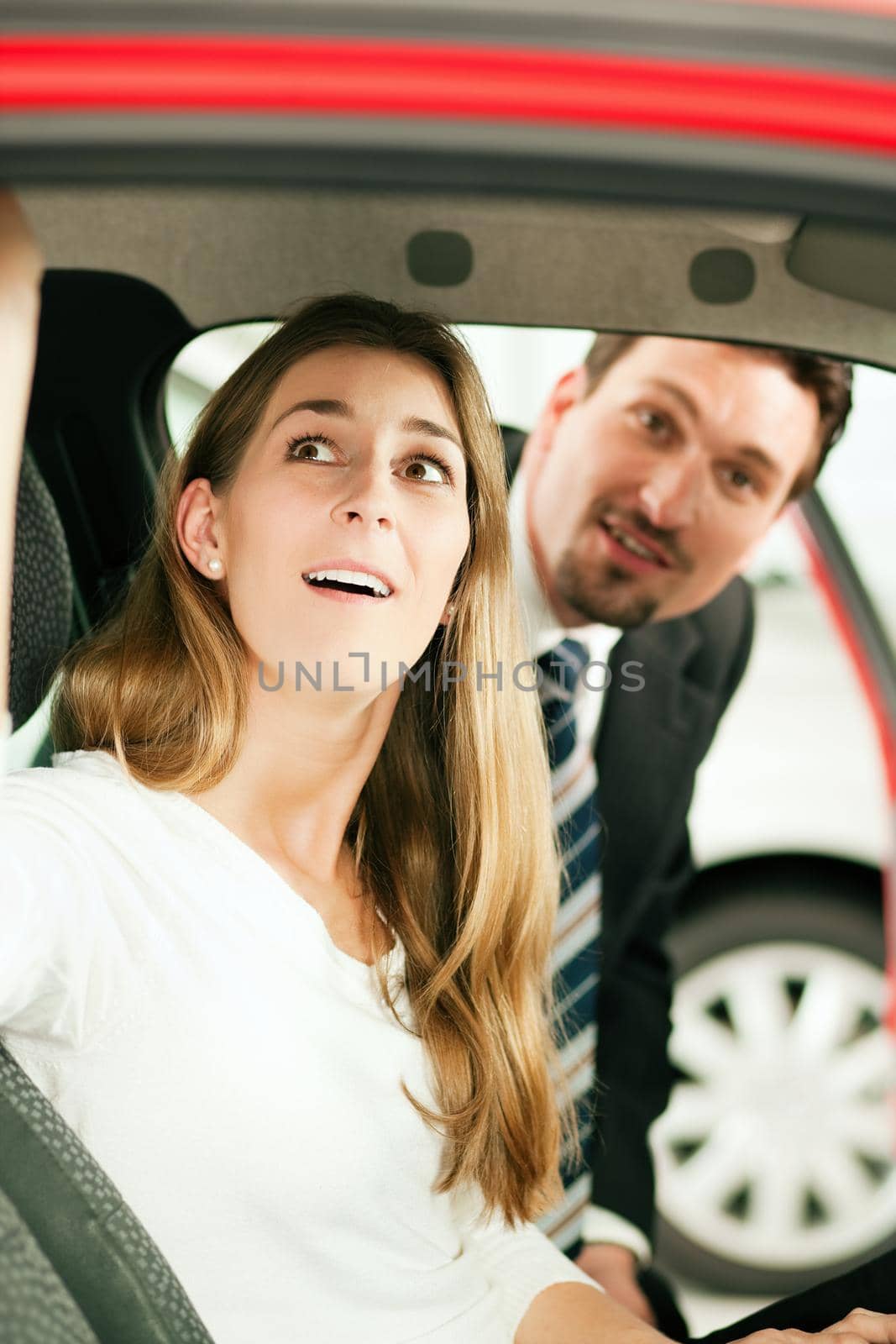 Woman buying a car in dealership sitting in her new auto, the salesman talking to her in the background