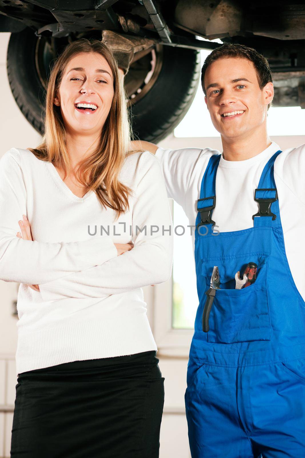 Woman standing in front of her car which is lifted on an auto hoist, a mechanic doing a repair underneath the auto