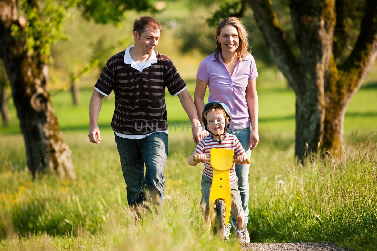 Family having a walk outdoors in summer, their little son using a training bike, unspoiled nature