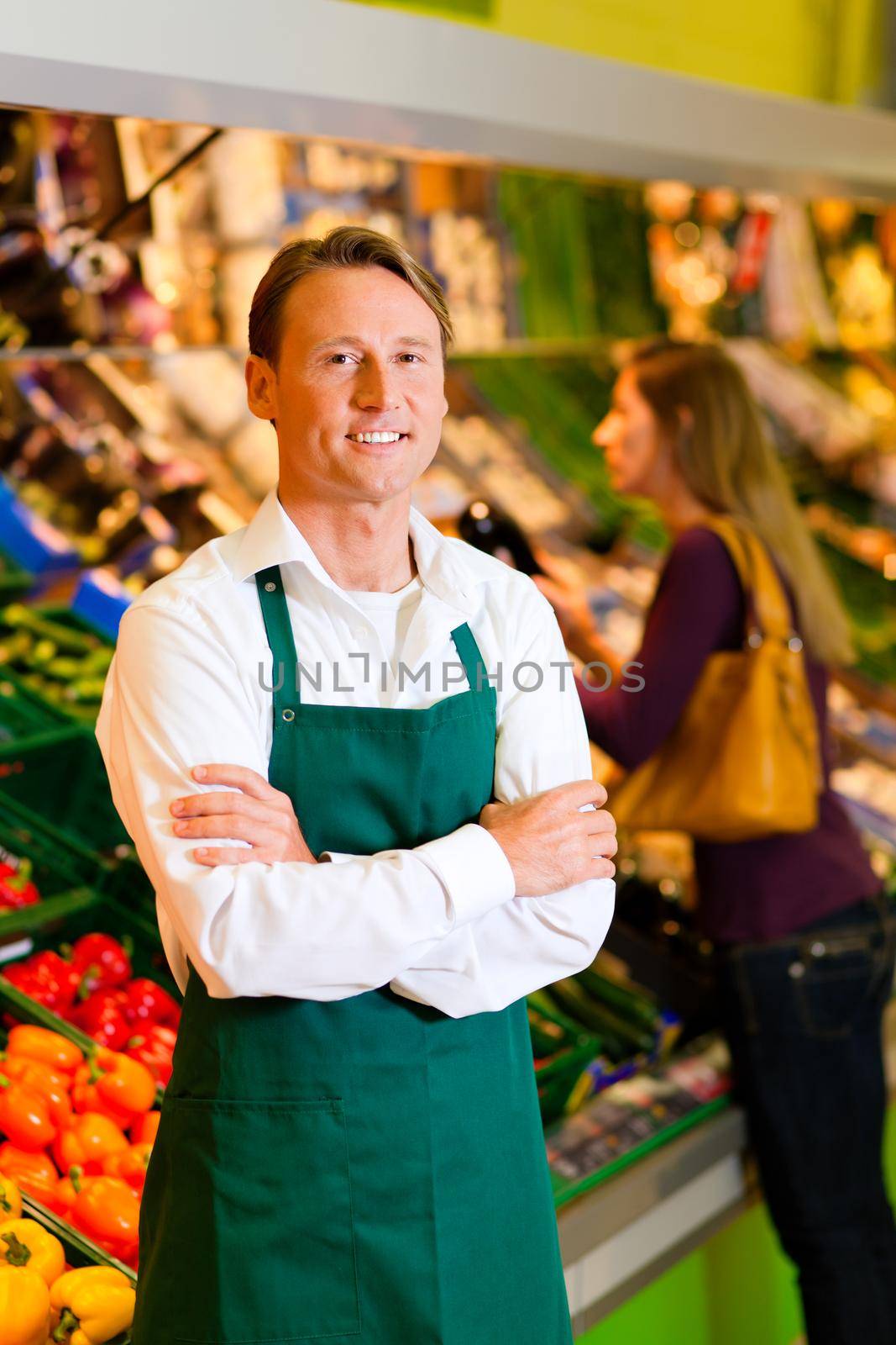 Shop assistant in a supermarket at the vegetable shelf; in the background a woman choosing vegetables