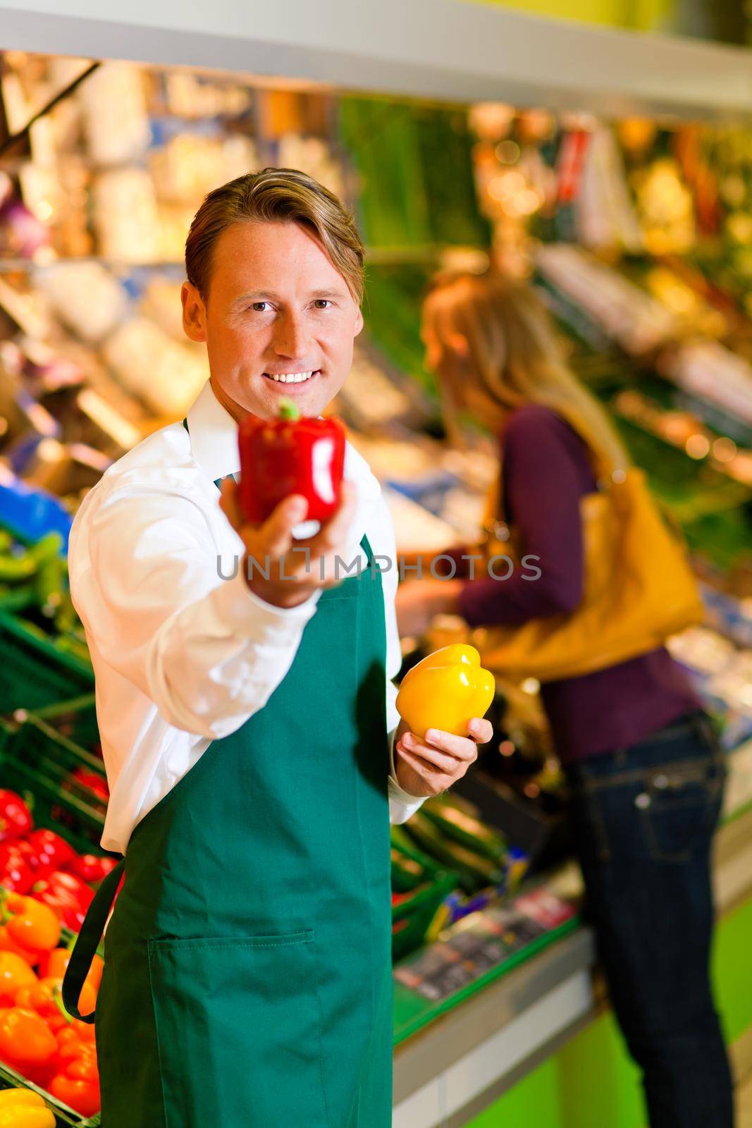 Shop assistant in a supermarket at the vegetable shelf; in the background a woman choosing vegetables