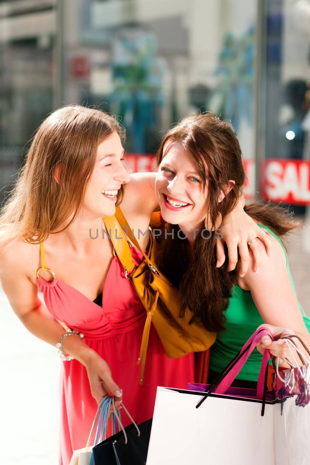 Two women being friends shopping downtown with colorful shopping bags, in the background a store can be seen with the words "sale" in the window