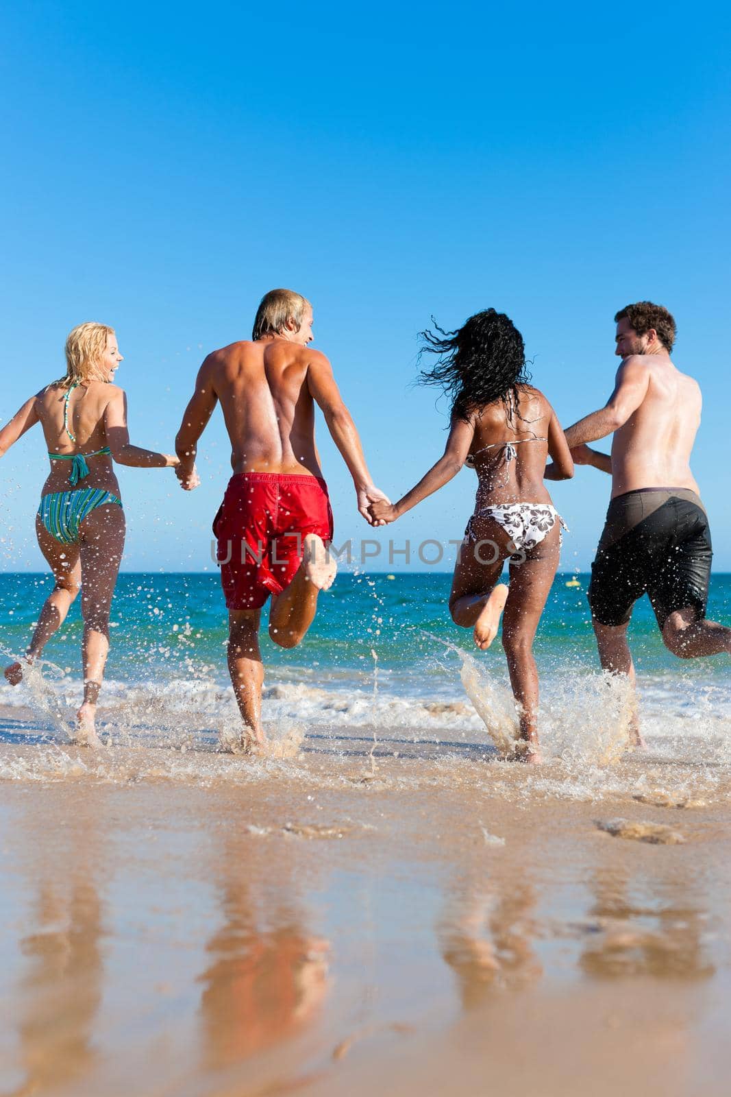 Four friends - men and women - on the beach having lots of fun in their vacation running through the water