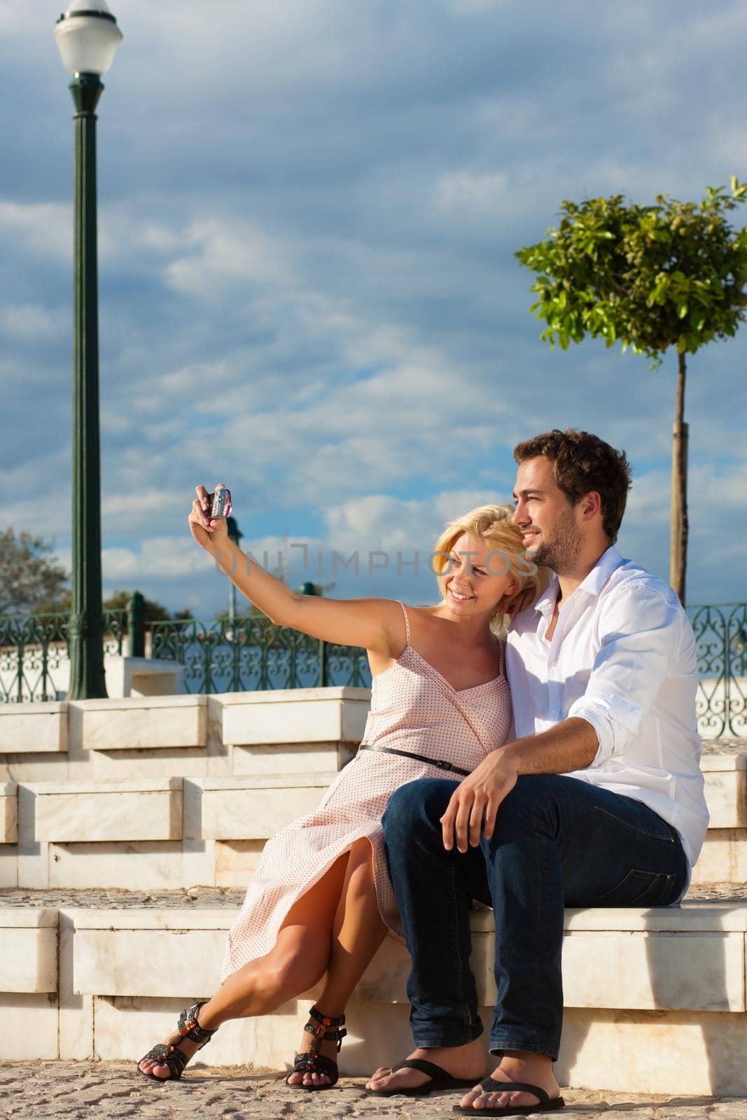 Couple having a city break in summer sitting on a brick wall in the sunlight, making pictures