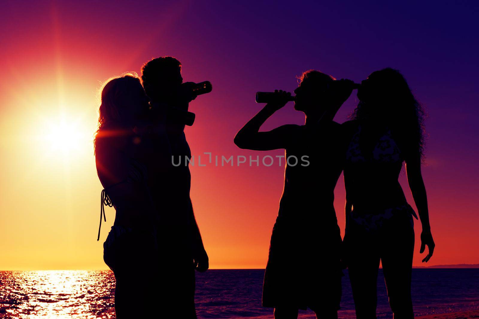 People (two couples) on the beach having a party, drinking and having a lot of fun in the sunset (only silhouette of people to be seen, people having bottles in their hands with the sun shining through)