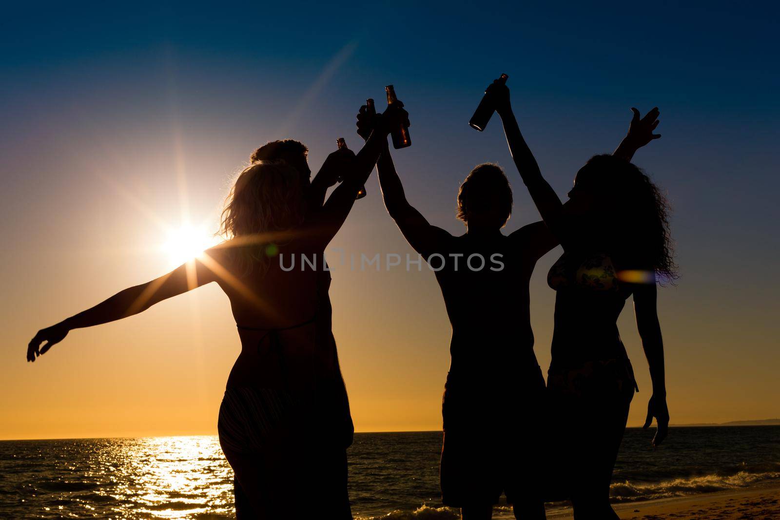 People (two couples) on the beach having a party, drinking and having a lot of fun in the sunset (only silhouette of people to be seen, people having bottles in their hands with the sun shining through)
