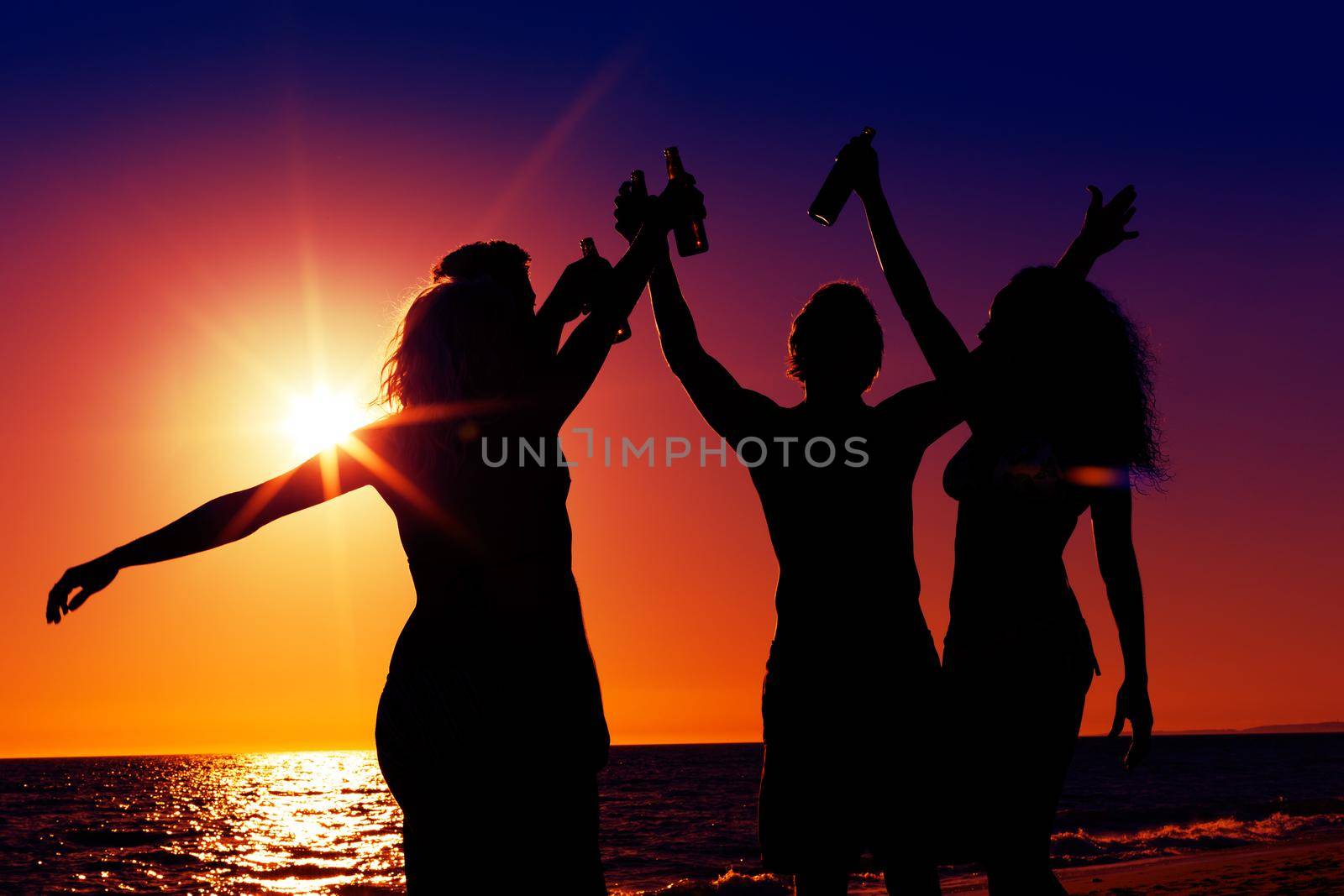 People (two couples) on the beach having a party, drinking and having a lot of fun in the sunset (only silhouette of people to be seen, people having bottles in their hands with the sun shining through)