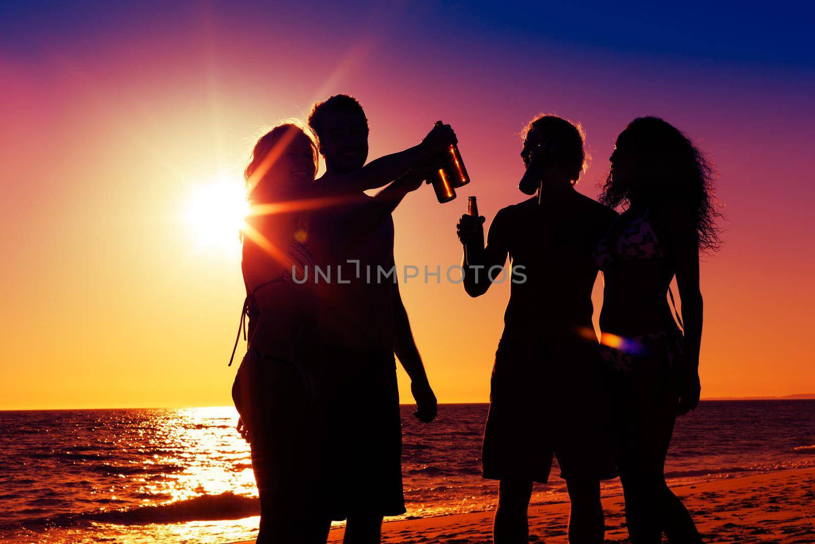 People (two couples) on the beach having a party, drinking and having a lot of fun in the sunset (only silhouette of people to be seen, people having bottles in their hands with the sun shining through)