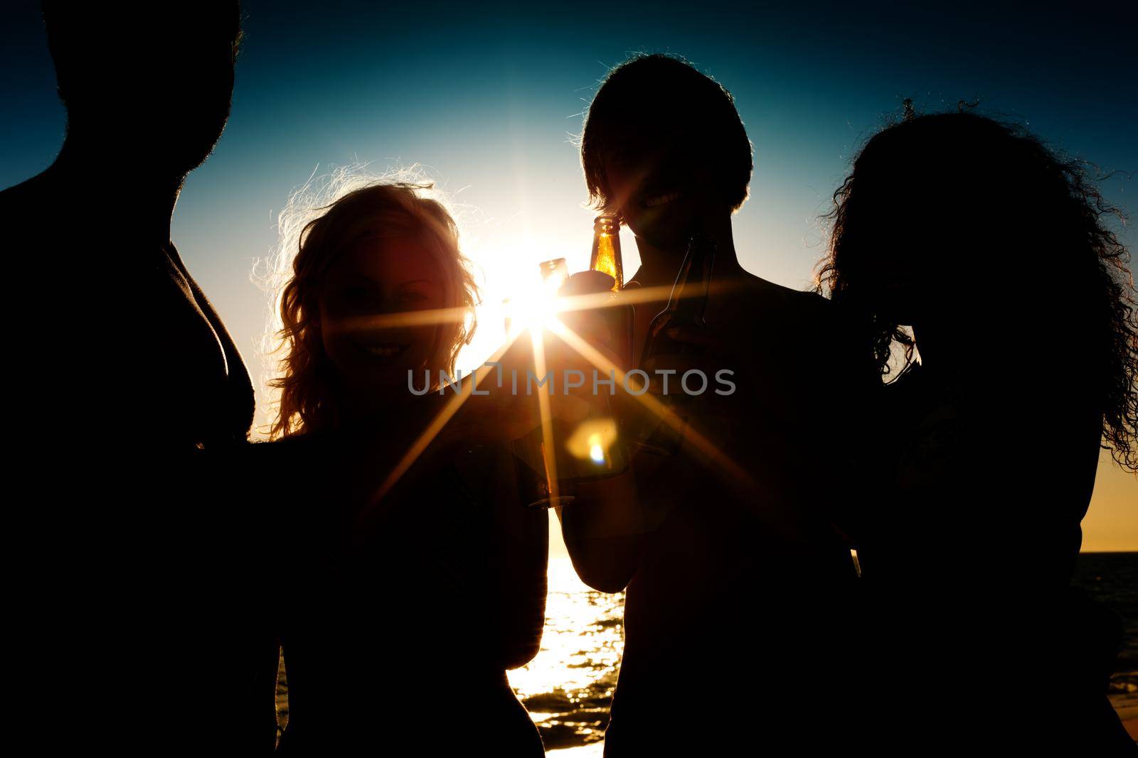 People (two couples) on the beach having a party, drinking and having a lot of fun in the sunset (only silhouette of people to be seen, people having bottles in their hands with the sun shining through)