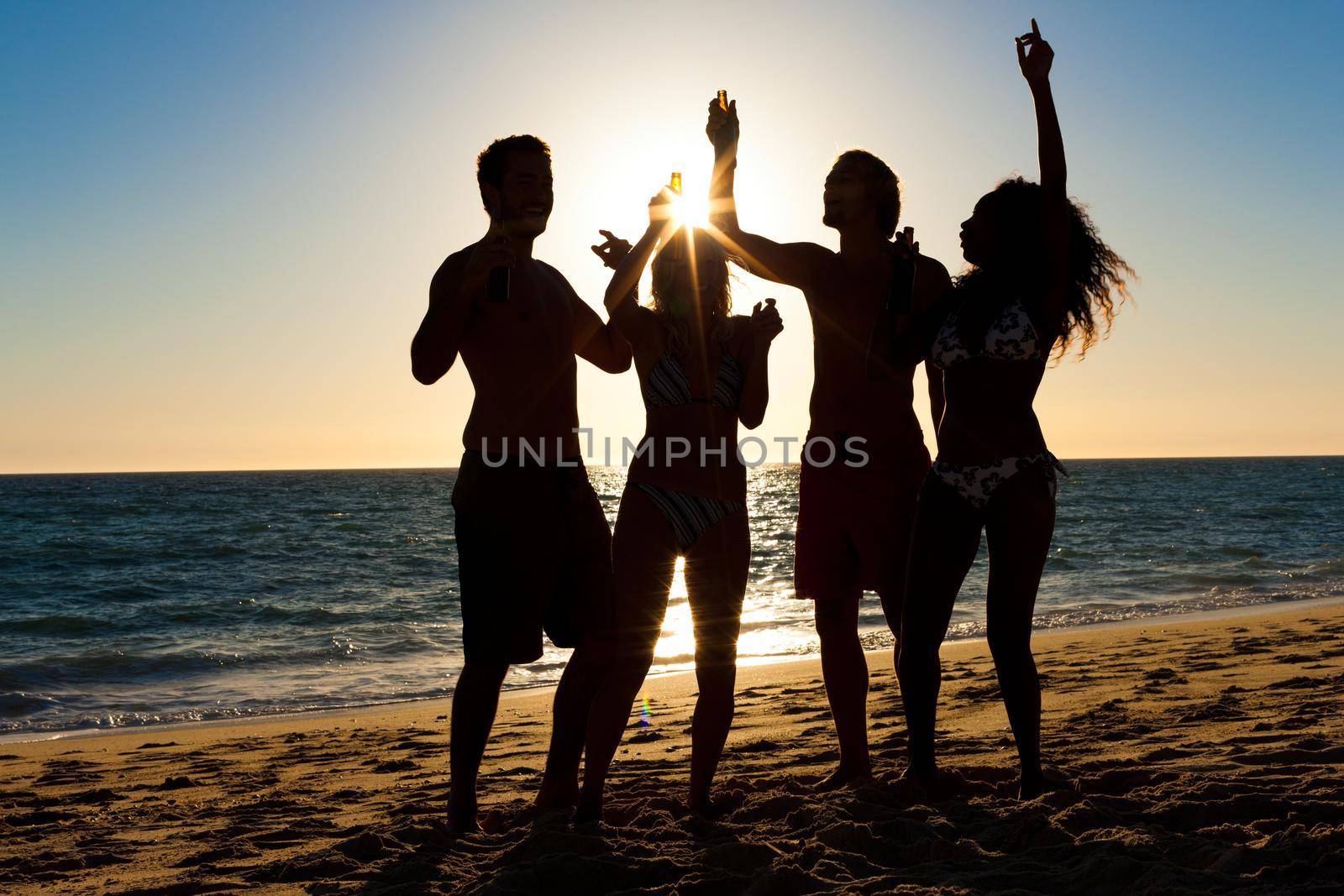 People (two couples) on the beach having a party, drinking and having a lot of fun in the sunset (only silhouette of people to be seen, people having bottles in their hands with the sun shining through)
