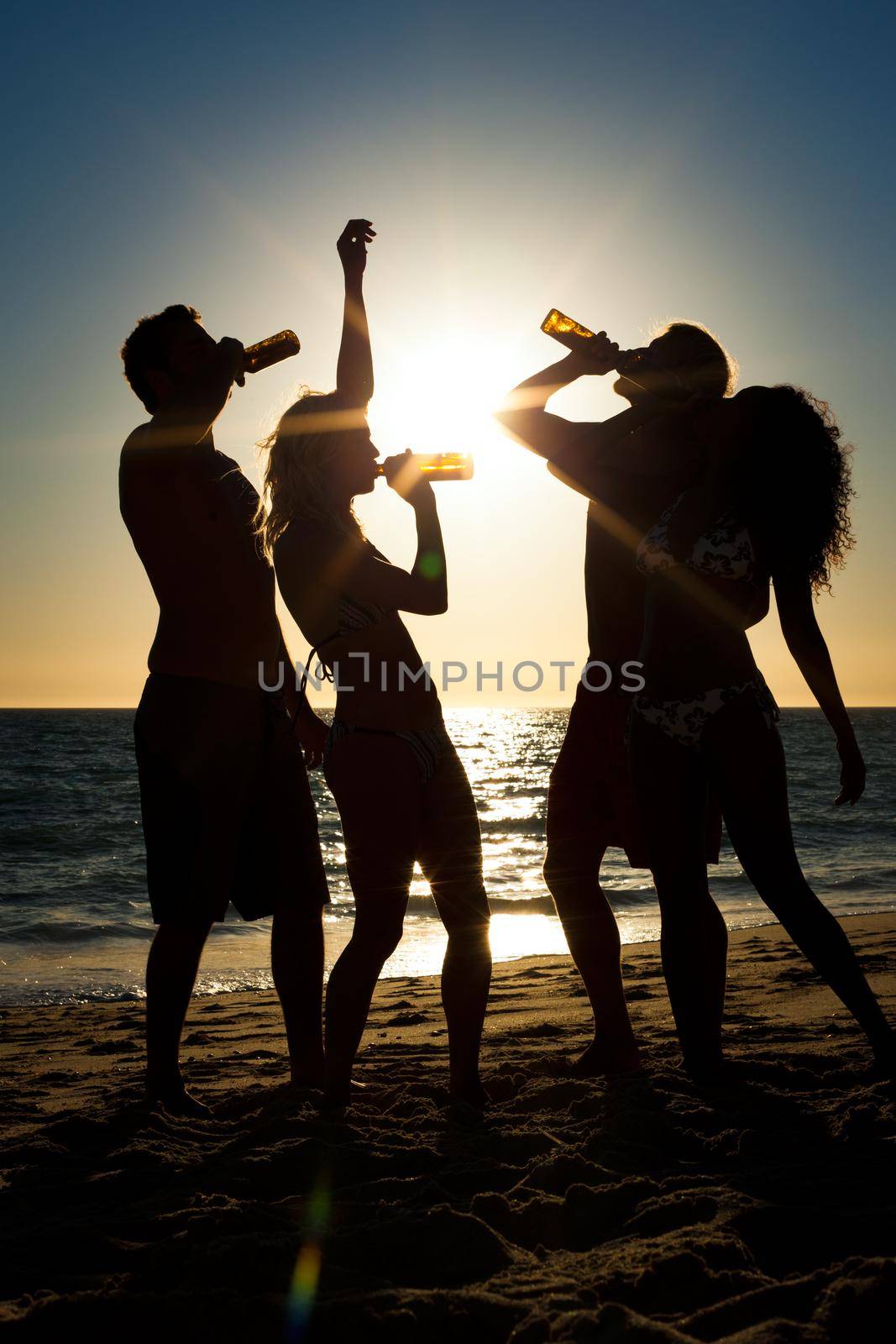 People (two couples) on the beach having a party, drinking and having a lot of fun in the sunset (only silhouette of people to be seen, people having bottles in their hands with the sun shining through)